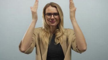 Beyaz bir arkaplan karşısında bej ceketli, gülümseyen sarışın kadın profesyonel bir ortamda neşe ve güven taşıyor..