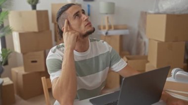 Dizüstü bilgisayarda parmakları klavyede yazan bir adam, taşınan kutular arasında taşınıyor, kulaklık yanında; odaklanma ayarlanıyor.