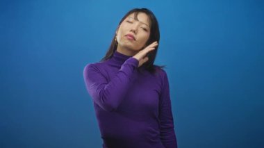 Stüdyoda elleri çenesinin altında mor balıkçı yaka giyen, mavi arka planda poz veren Asyalı genç bir kadın..
