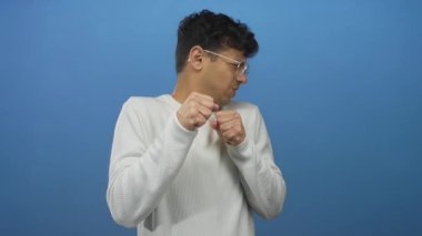 Yumruklarını sıkmış ve gözlüklü bir adam canlı mavi bir arkaplan karşısında stüdyoda duruyor..