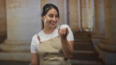 Kadın, taş sütunlarla binada el hareketi yaparken görülüyor; dostça bir davet..