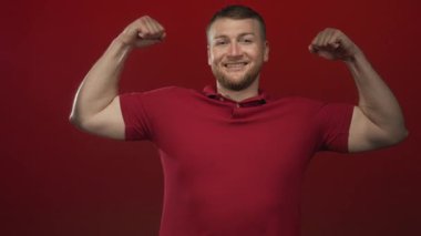 Genç, tıknaz bir adam, kırmızı bir stüdyo setinde kaslarını sıkmış, objektife gülümsüyor, özgüven kuvveti.
