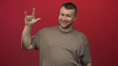 Stüdyo kırmızı duvarında, sakallı ve günlük bej tişörtlü gülümseyen, sağ elini kaldırmış kaya boynuzlarını ve görünür avuç içini gösteren adam; neşeli enerji.