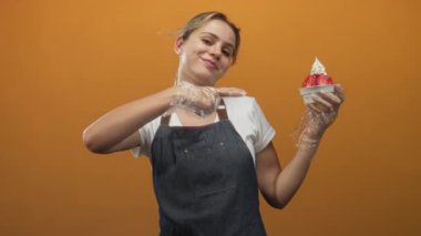 Kadın, turuncu stüdyoda kot önlük ve temiz eldivenler giyerken çilekli dondurmayı ve bir fincan yumuşak şekeri işaret ediyor..