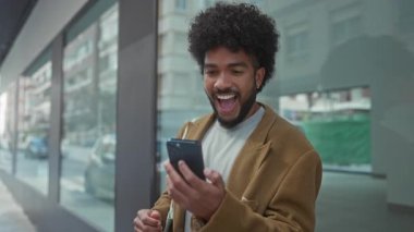 Şehir merkezinde akıllı telefondan başarıyı kutlayan bir adam şehir merkezinde neşeli bir ifadeyle sıradan bir ceket giyiyor ve geniş bir gülümseme sergiliyor..