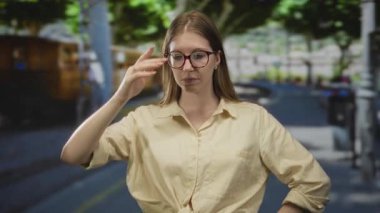 Gözlüklü ve bej tişörtlü bir kadın yeşil ağaçların altında elleri ile alnına dokunuyor..