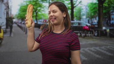 Kadın sokağın ilerisinde, park halindeki arabaların yanında kameraya gülümseyen bir baget tutuyor ve işaret ediyor..