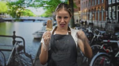 Bisikletleri ve binaları görünür halde olan Amsterdam Kanalı 'nın yanındaki bir sokakta buzlu şeker tutan ve kulağına yumruk yapan bir kadın..
