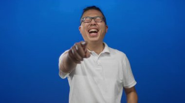 Mavi stüdyoda beyaz polo giyen bir adam iki parmağıyla işaret ediyor ve görünür dişleriyle gülüyor..