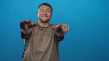 Genç sakallı adam gülümsüyor ve parlak mavi stüdyoda iki elini de kameraya doğrultuyor, kahverengi bir tişört ve rahat bir duruş giyiyor; dostça bir davet.