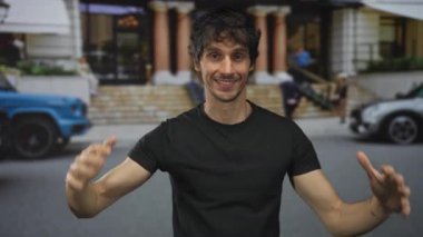 Genç adam hareketli caddede sütunlu binanın önünde dururken enerjik bir gülümseme ile el sallıyor; sıcak neşeli karşılama.