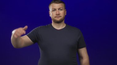 Koyu mavi bir stüdyoda çıplak kollu ve tişörtlü adam el pençe divan selam veriyor; görev saygısı.