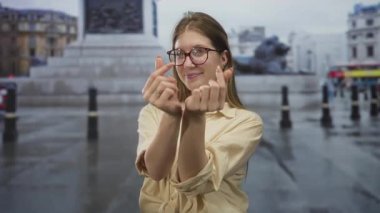 Kadın, yağmurlu caddelerde, taştan heykeller ve şehir binalarıyla parmak kalp hareketi sergiliyor..
