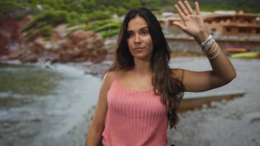 Kadın, plajda el kaldırır ve pembe kolsuz tişörtle avuç içi ve çıplak omuz gösterir; sakin yansıma.