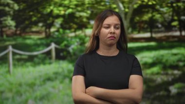 İp çitin yanında kolları çaprazlanmış bir kadın ve güneş ışığı altında ağaçlar, siyah bir tişört giyerken ince bir sırıtışla yukarı bakıyor; sinir bozucu.