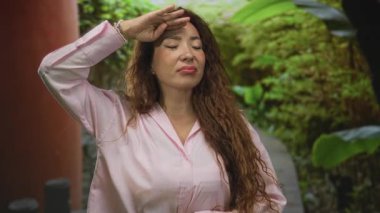 Gülümseyen ve bakan, pembe bir gömlek ve uzun kıvırcık saçlar giyen, gür bir bahçede eli ile yüzünü koruyan kadın; mutluluk özgüveni.