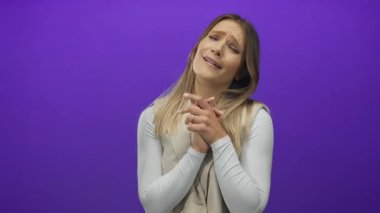 Sarışın, mor arka planda gülümseyen bir kadın, kapalı elleriyle duygularını ifade ediyor, günlük beyaz kıyafetler giyiyor, mor duvarın üzerinde duruyor..