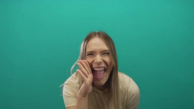 Genç sarışın kadın yeşil arka planda kameraya doğru sevinçle bağırıyor. Heyecan ve coşku ifadelerini vurguluyor..