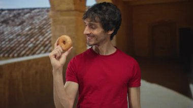 Kırmızı gömlekli genç adam gülümsüyor ve ağızdan ağıza jöleli donut tutuyor. Tarihi üniversite binası girişinin önünde ısırıyor. Açlıktan zevk alıyor..