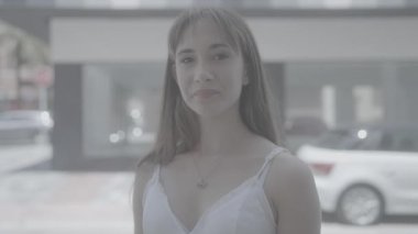 Gülümseyen genç kadın, elleri sokakta, park halindeki arabanın yanında beyaz elbiseyle kalp yapıyor..