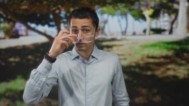 Genç İspanyol bir adam güneşli bir parkta gözlük takıyor. Etrafında ağaçlar ve çimenler var. Rahat bir açık hava yayıyor..