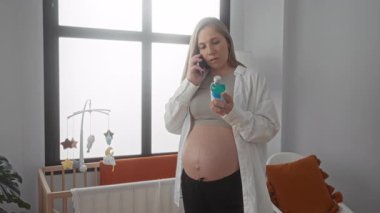 Sarı saçlı hamile bir kadın, elinde beşikli bir bebek odasında şişeyle telefonla konuşuyor. Ev hayatını ve doğum hazırlıklarını ön plana çıkarıyor..