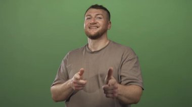 Adam gülümsüyor ve iki eli de yeşil stüdyo ortamında görülebilen kamerayı işaret ediyor; kendine güven yaklaşımı.