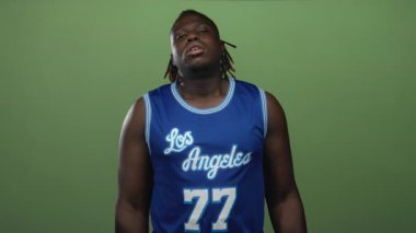 Mavi Los Angeles basketbol forması giyen adam 77 numaralı formayı giymiş. Stüdyoda yeşil arka planda görünen çıplak kollar ve rahat omuzlarla yukarı bakıyor..