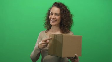 Yeşil bir stüdyoda pakete işaret ederken gülümseyen ve elinde karton bir kutu tutan bir kadın..