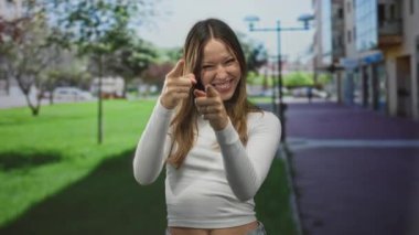 Woman points fingers directly at camera on city street, smiling, wearing white crop top and showing bare midriff; playful confidence.