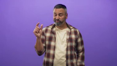 Gri sakallı ve kısa gri saçlı bir adam. Mor bir stüdyoda başparmak ve işaret parmağı sıkıyor. Ekose gömlek giyerken..