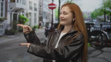Kızıl saçlı kadın başparmağıyla işaret ediyor ve işaret parmağıyla işaret ediyor, çıplak elle görülüyor, sokakta deri ceket giyiyor, neşeli bir cazibesi var..