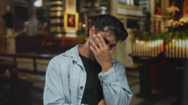 Kotlu tişörtlü bir adam, bina içinde yıpranmış bir kilise sırasının ve sıralanmış mumların yanında yanak yanağa gülümsüyor..