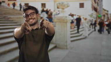 Adam binanın ön cephesindeki kalabalık sokak merdivenlerinde kamerayı işaret ediyor, gülümsüyor ve gözlük takıyor; neşeli bir davet.