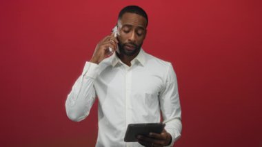 Adam telefonu kulağına tutuyor ve stüdyoda kırmızı arkaplan ile tablet okuyor; odaklanmış verimlilik uzaktan çalışma.