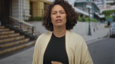 Kadın, şehir caddesinde, örgü hırka giyerken taş bina merdivenlerinin yanında, kapalı gözlerle omzuna dokunuyor..