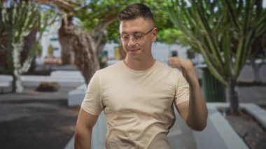 Gözlüklü bir adam ellerini başının arkasına koyarak gülümsüyor, kaktüs ağaçlarıyla dolu bir sokakta bej tişört giyiyor; rahatlamış özgüven..