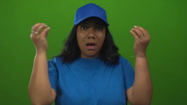 Teslimatçı kadın yeşil stüdyoda mavi şapka ve gömlek giyerek omuz silkerek iki elini kaldırıyor..