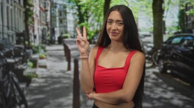 Park halindeki arabalar ve bisikletlerle ağaçlık bir caddede el işaretini gösteren genç bir kadın, neşeli bir güven..