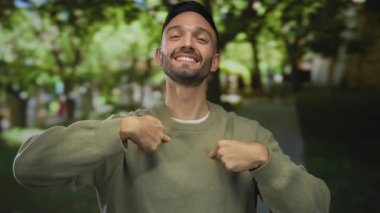 Sakallı genç bir adam yeşil arka planda gülümsüyor, günlük kıyafetler giyiyor, kendine güvenli bir şekilde açık havada, mutluluğu ve pozitifliği simgeliyor..