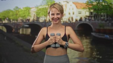 Kanal köprüsü ve spor sutyeni takıp spor yaparken gülümseyen, spor yaparken gülümseyen, elleri göğsüne kadar kanala dayalı halterler tutan sarışın kadın..