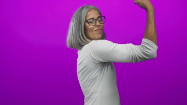 Gri saçlı bir kadın, mor stüdyoda kolunu esnetirken çıplak pazılarını gösteriyor..