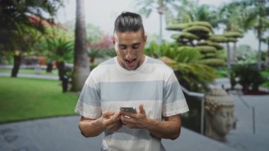 Akıllı telefon tutan, başparmaklarını tıklatan ve binanın dışındaki bir bahçedeki Buda heykelinin yanına bakan adam, odaklanmış bağlantı.