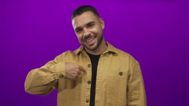 Bronz ceket giyen adam gülümsüyor ve mor stüdyoda başparmağını kaldırırken parmağıyla gömleğini işaret ediyor..