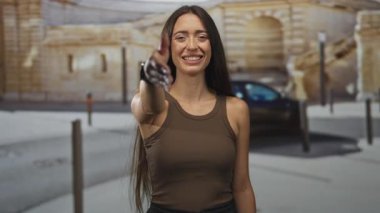 Kadın, park halindeki araba ve taş bina yakınında el sıkışmak için sağ elini uzatıyor, kapalı gözlerle gülümsüyor; selamlaşmayı karşılıyor.