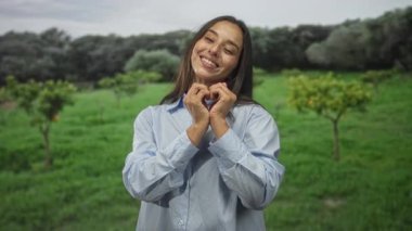 Gülümseyen kadın, elleri yeşil bir alanda ağaçlar ve otlarla dolu bir kalp oluşturur; sevgi bağı topluluğu.