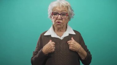 Üst düzey kadın göğsünü işaret ediyor ve stüdyoda doğrudan kameraya konuşurken kazak yakasını düzeltiyor..