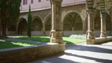 Kavisli manastır avlusu, kemerler ve sütunlar, güneşli taş kaldırımlar ve yumuşak bokeh bahçesinde çimenler; arka plan sakin.