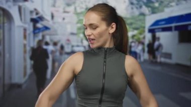 Kadın, yeşil kolsuz yelek giyerken çıplak kol kaslarıyla halteri kaldırıyor ve sokakta gülümsüyor..