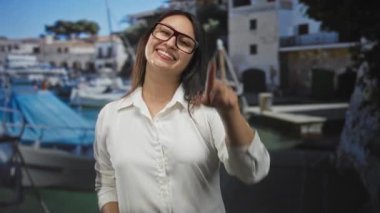 Genç İspanyol kadın deniz kenarı rıhtımında tekneleri olan bir kamerayı işaret ediyor..